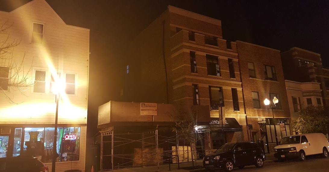 Straßenansicht bei Nacht mit Baugerüsten vor einem Geschäftsraum in einem Backsteingebäude, was auf Renovierungsarbeiten in einem städtischen, kommerziellen Bereich hindeutet.